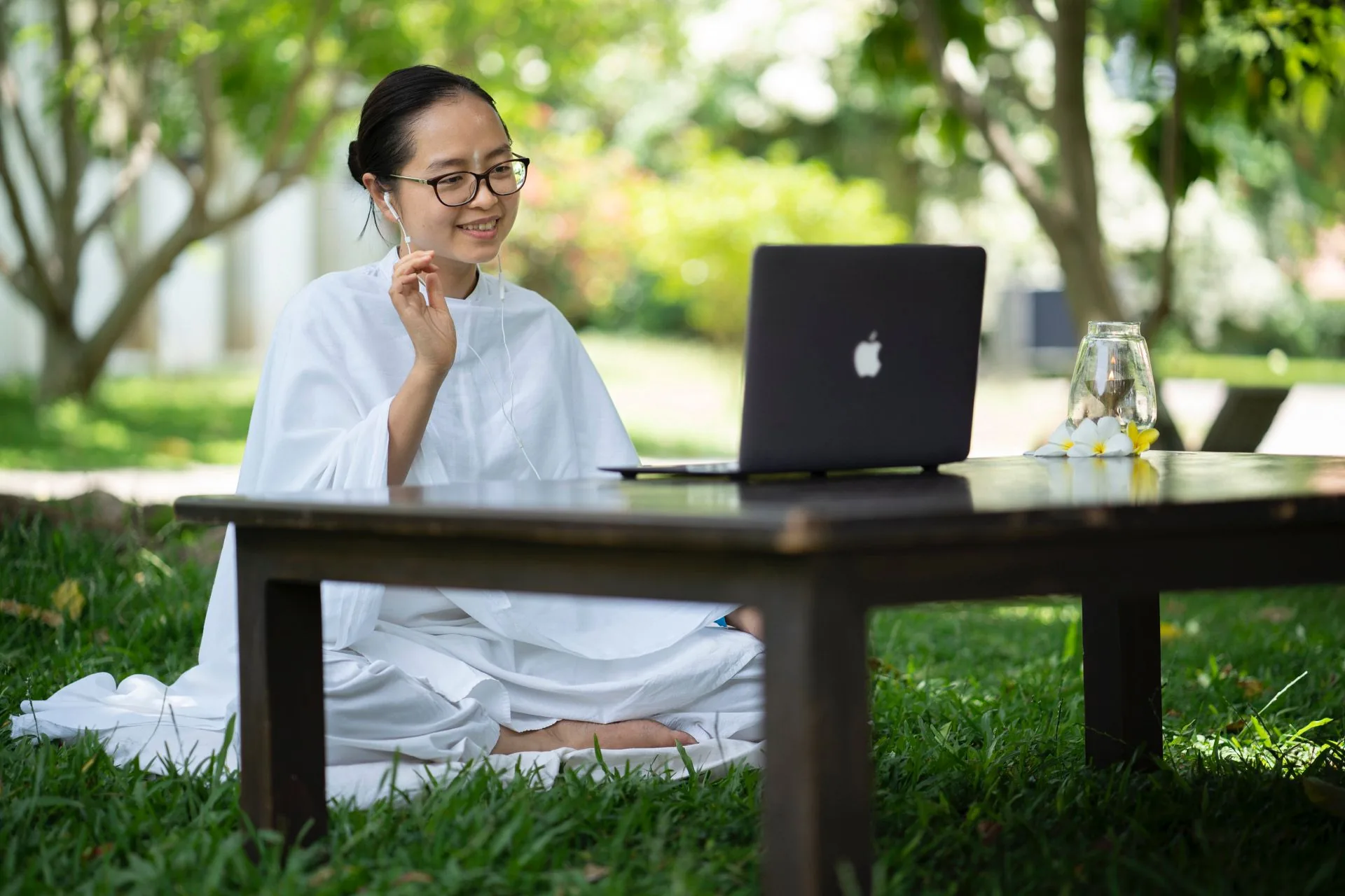 Frau in weißer Robe sitzt im Garten im Schneidersitz vor einem Laptop auf einem Holztisch