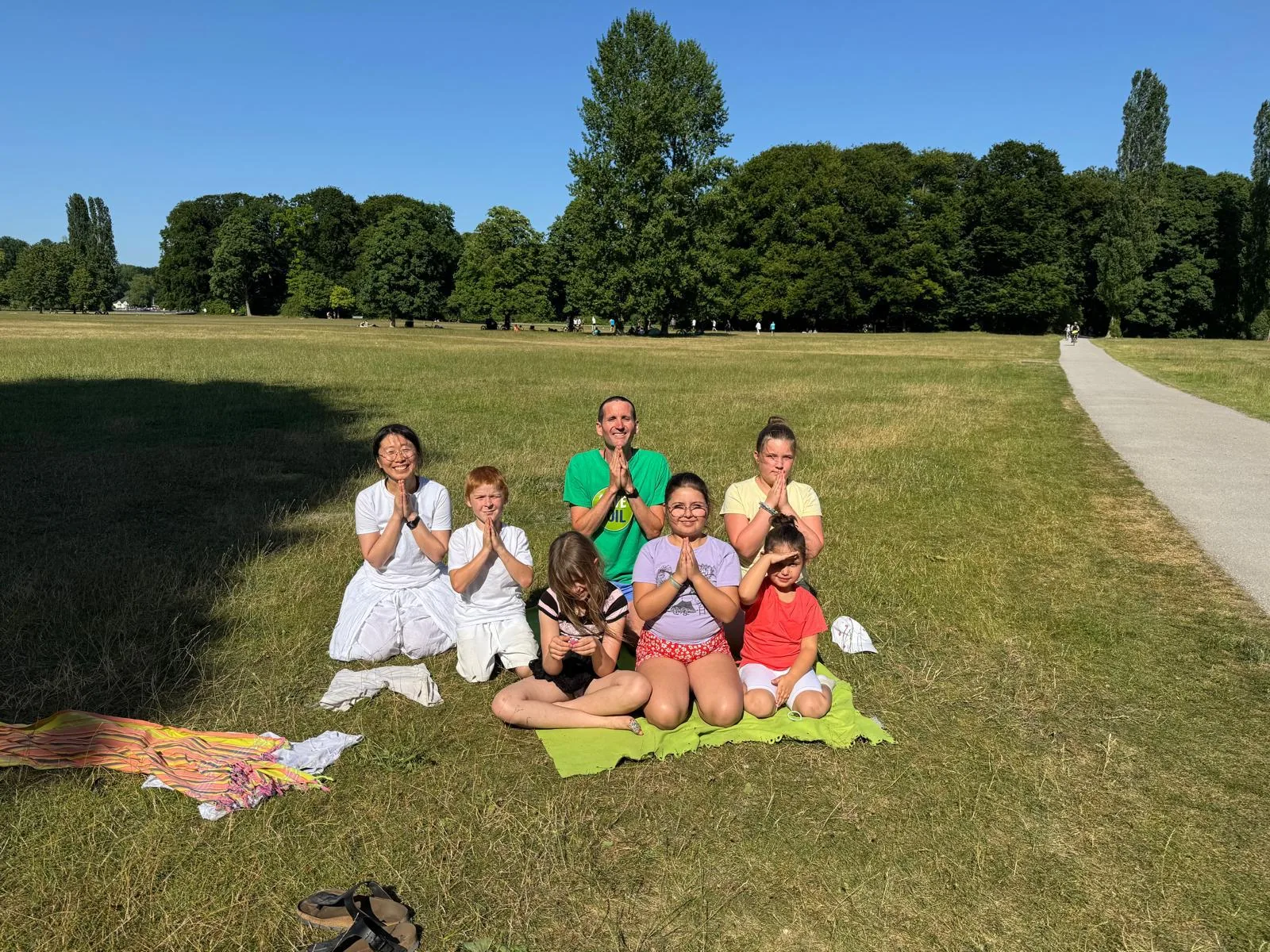 Lehrerin und sechs Kinder sitzen im Namaste-Gruß auf einer sonnigen Wiese