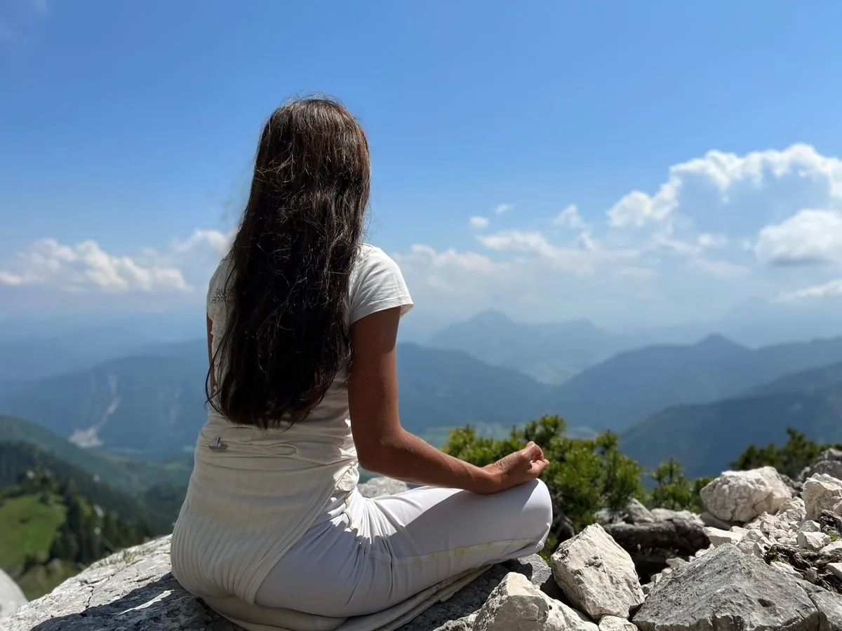 Frau meditiert im Schneidersitz auf einem Gipfelfelsen über einer Berglandschaft